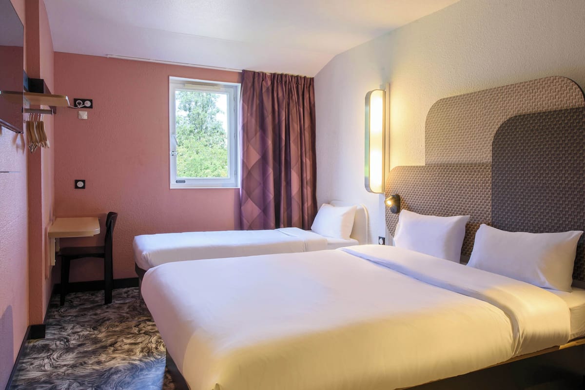 A hotel room with two beds, a desk, a chair, a window with curtains, and a wall-mounted lamp.
