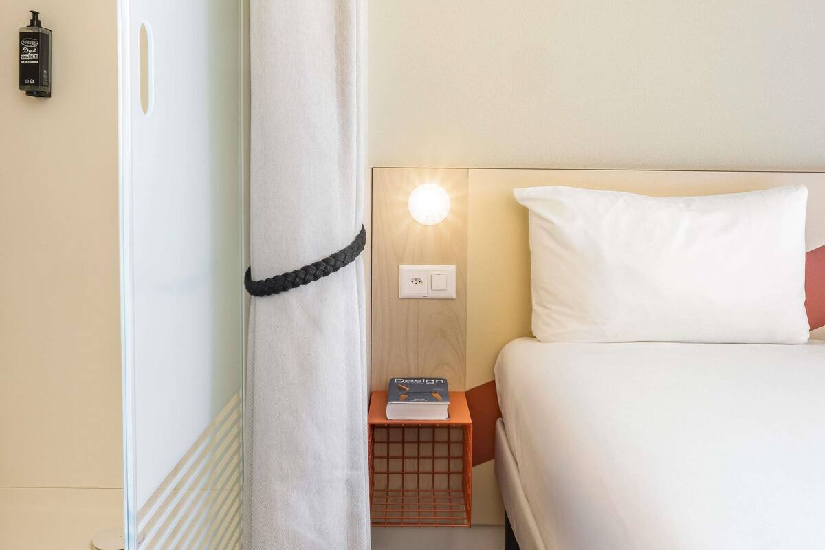 A hotel room with a bed, a nightstand, a book, a lamp, and a wall-mounted phone.
