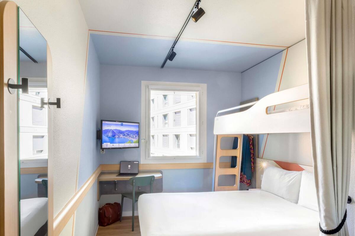 A compact room with a bunk bed, a desk with a laptop, a television, and a window with blinds.