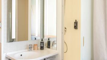 Une salle de bain moderne avec un lavabo blanc, un miroir et une douche. On y trouve des articles de toilette sur le lavabo et un porte-serviettes.