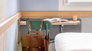 Een hoek van een kamer met een groene stoel, een bruine lederen tas, een houten bureau met een boek en een beker, en een wit bed.
