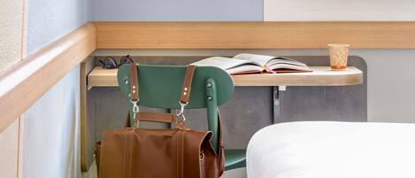 A corner of a room with a green chair, a brown leather bag, a wooden desk with a book and a cup, and a white bed.