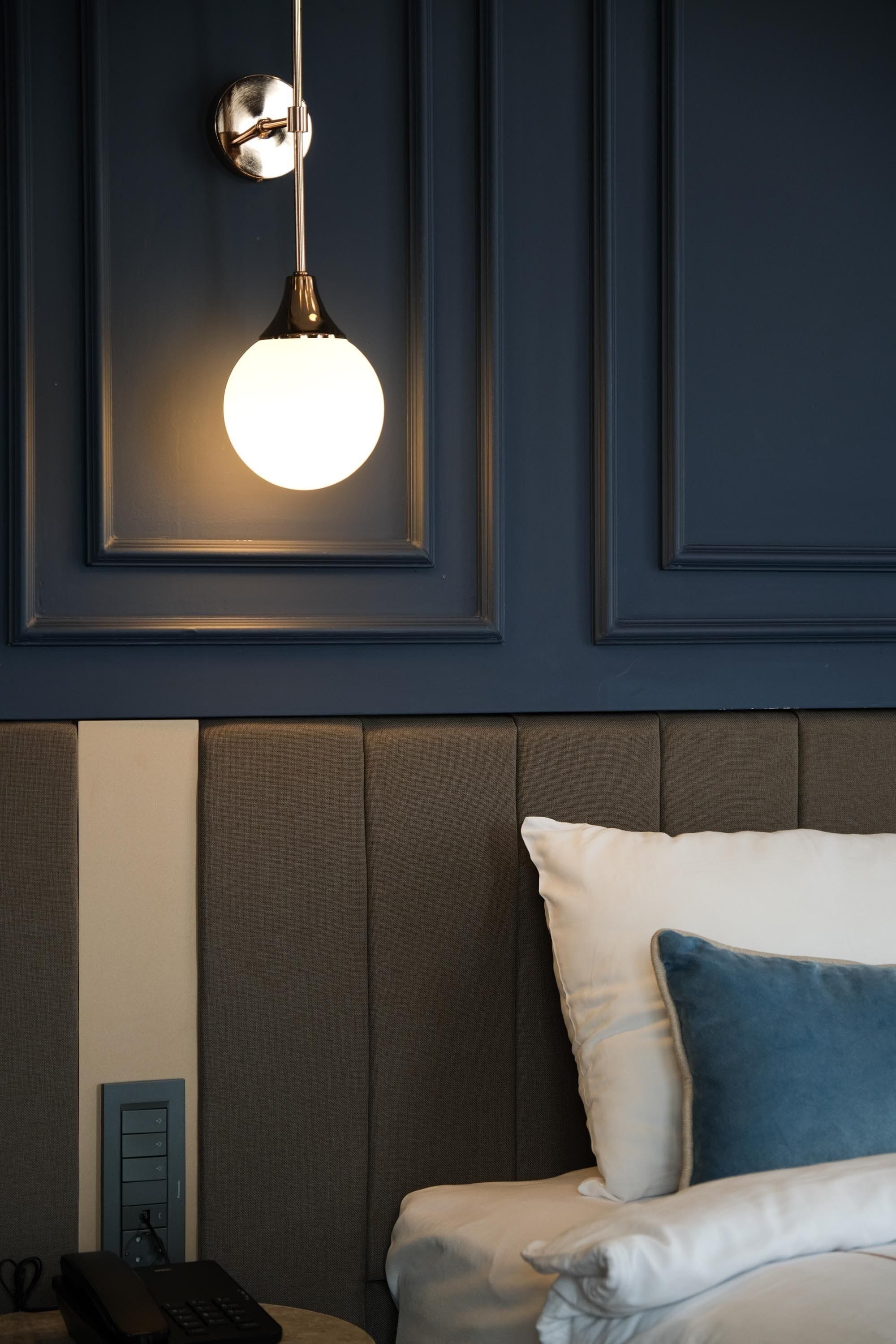 Habitación de hotel con una pared azul oscuro, una lámpara redonda, una cama con almohadas blancas y azules, y una mesita de noche con teléfono y control remoto.