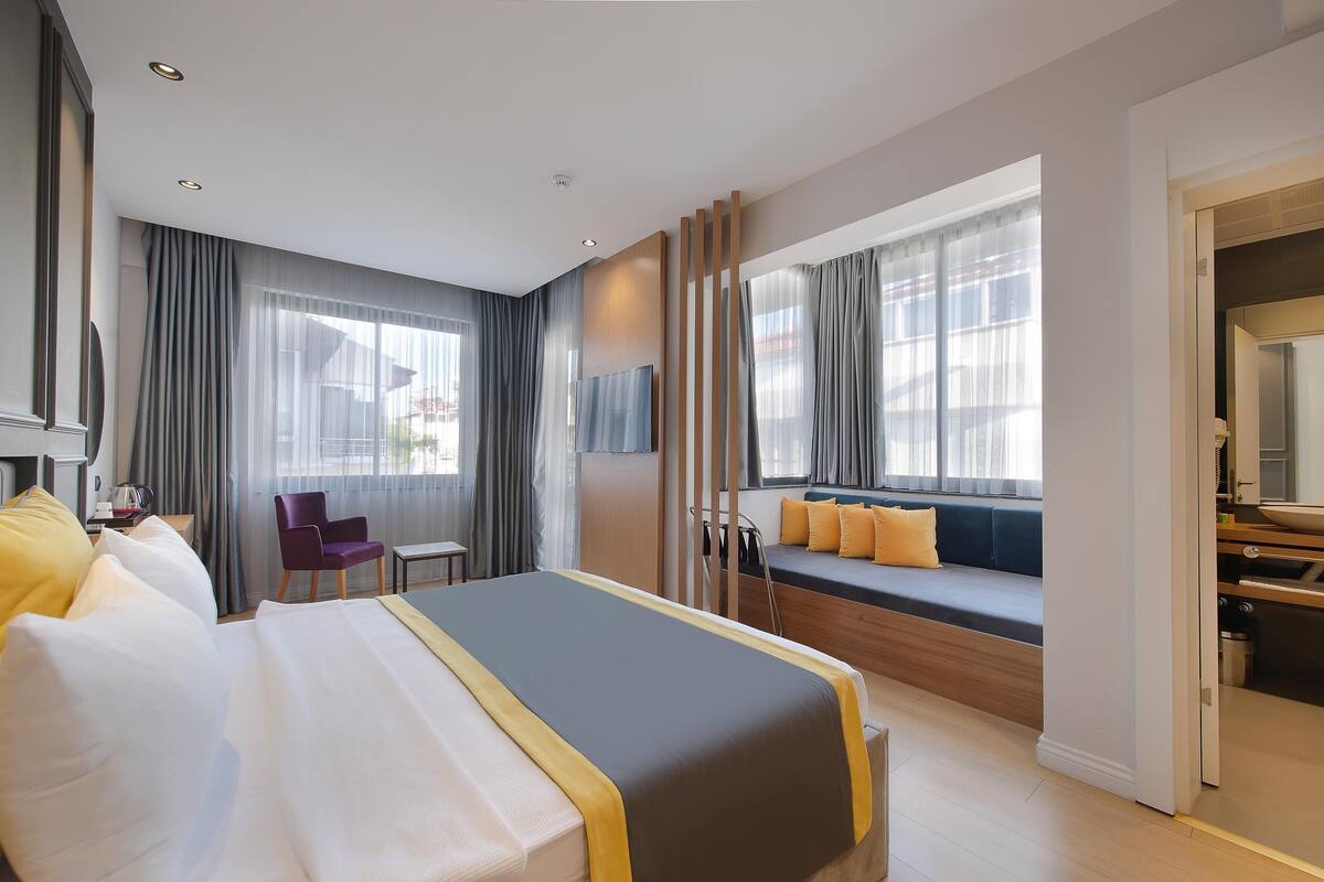 Une chambre d’hôtel moderne dotée d’un grand lit, d’un coin salon avec des coussins, d’un bureau et offrant une vue sur la ville grâce à de grandes fenêtres.