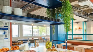 Una barra de desayuno con diferentes alimentos, una zona de estar con mesas y sillas, y vistas al exterior a través de amplios ventanales.