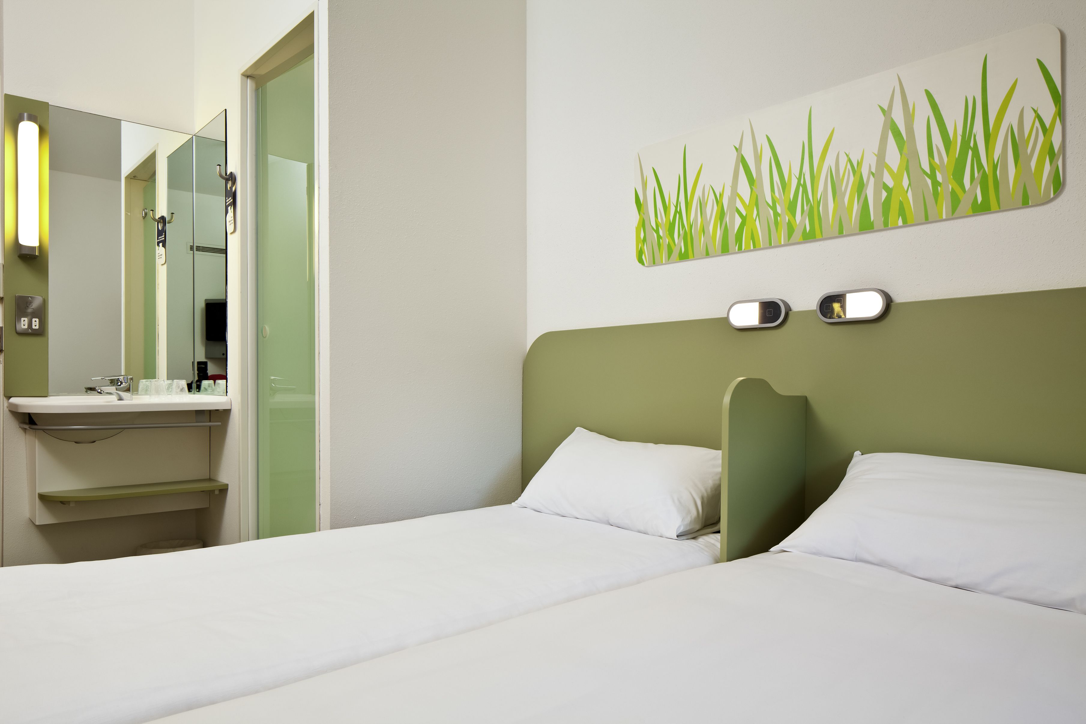 A hotel room with a bed, a green headboard, a wall-mounted light fixture, and a bathroom with a mirror and sink.