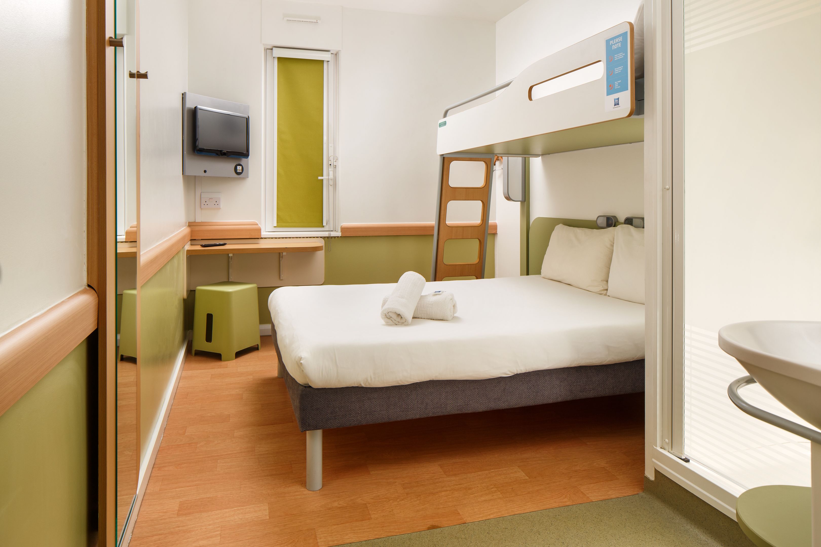 A compact hotel room with a bunk bed, a small desk with a television, a bathroom with a sink, and a mirror.