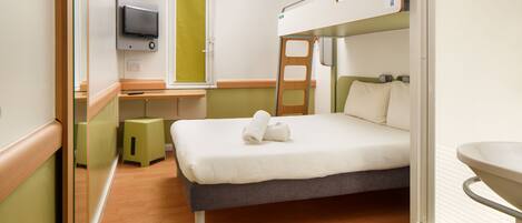 A compact hotel room with a bunk bed, a small desk with a television, a bathroom with a sink, and a mirror.