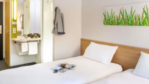 Habitación de hotel con cama, mesita de noche, espejo, baño con lavamanos y toallero, y una planta decorativa en la pared.