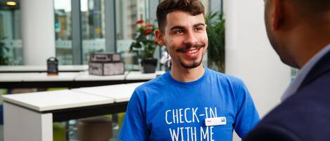 A person in a blue "Check-In With Me" t-shirt holding a smartphone, standing in an indoor setting with a modern interior, including a table and potted plants.