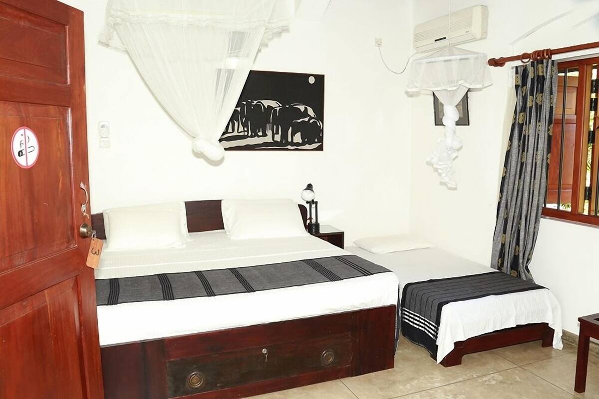 A bedroom with a large bed, a smaller bed, a nightstand, a bedside table, a wall-mounted air conditioner, a window with curtains, and a door with a lock.
