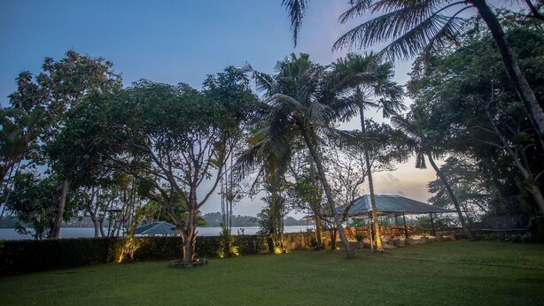 A serene outdoor area with a grassy lawn, palm trees, and a gazebo with a roof. - Serenity Lake Front Villa (Moratuwa)