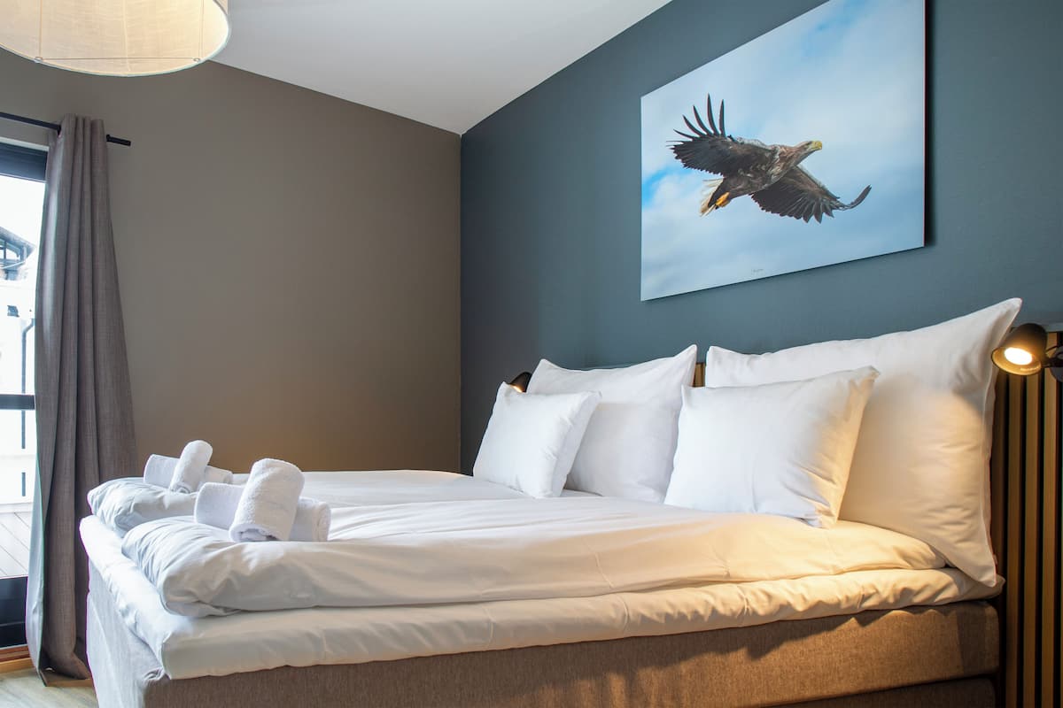 Una cama doble con sábanas y almohadas blancas, un ventanal con cortinas y un cuadro de un águila colgado en la pared.