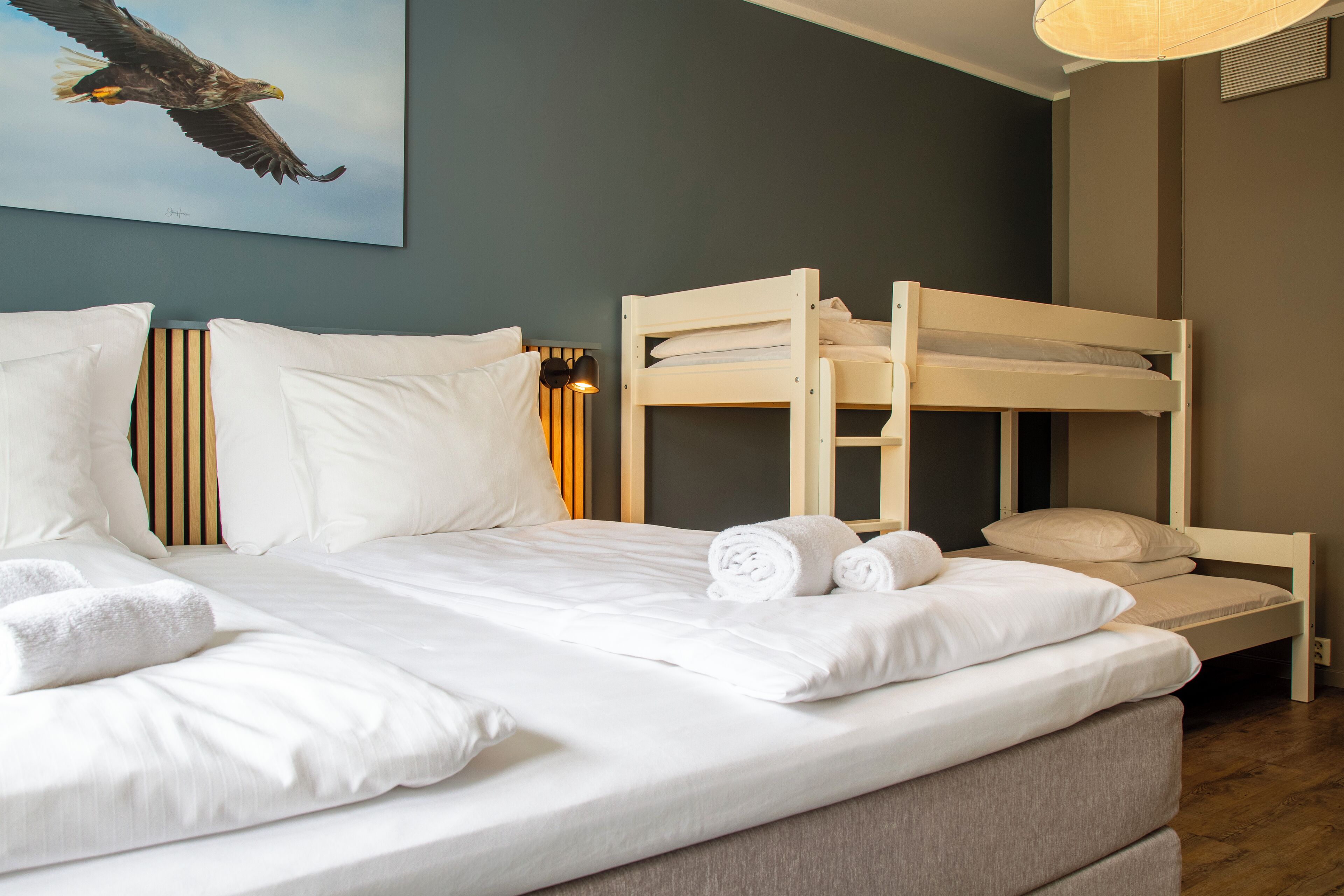 A neatly made bed with white linens and rolled towels, a bunk bed, and a painting of an eagle on the wall.