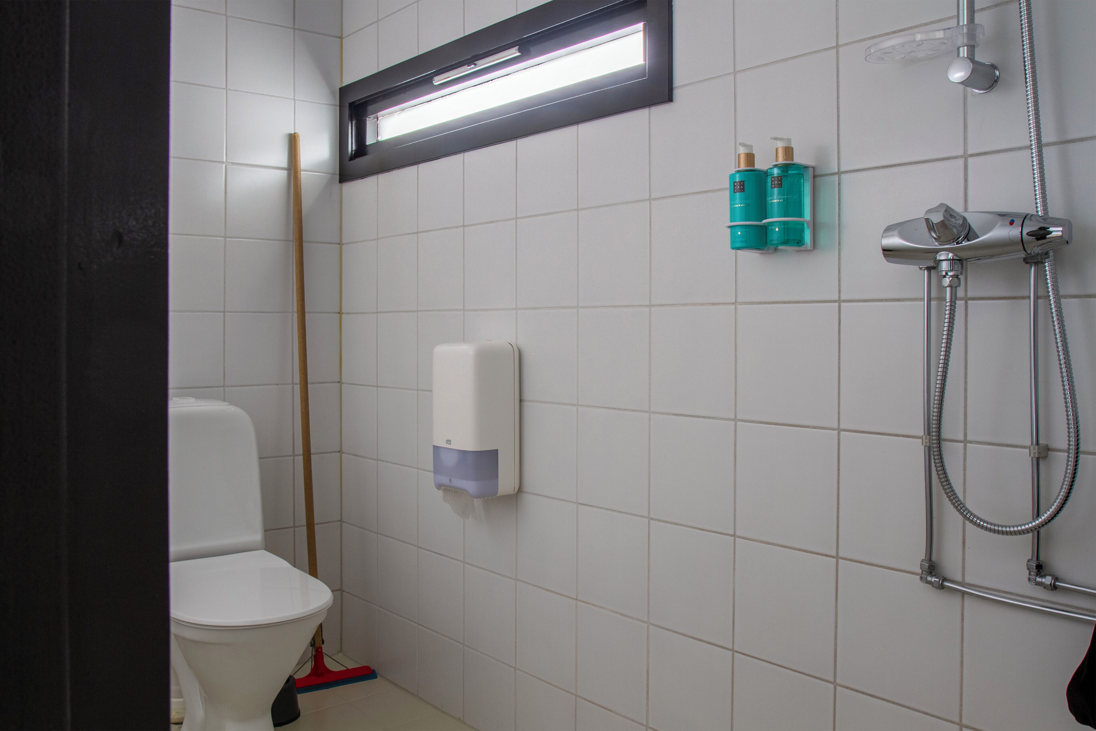 A bathroom with a toilet, a hand dryer, a shower with a handheld showerhead, and two bottles of toiletries on the wall.