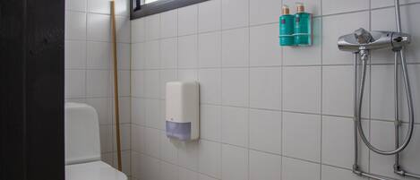 Une salle de bain équipée de toilettes, d’un sèche-mains, d’une douche avec un pommeau de douche amovible, et de deux flacons de produits de toilette fixés au mur.