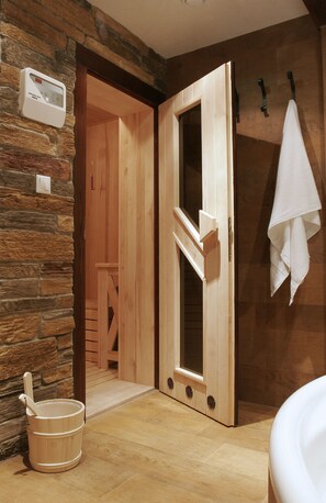 Una sauna de madera con pared de piedra, una puerta de madera, una toalla blanca colgada y un cubo de madera.