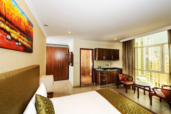 A hotel room with a bed, a dining area with chairs, a kitchen area, and a large window with a view of buildings. - Bravo Royal Hotel Suite (Salmiya)