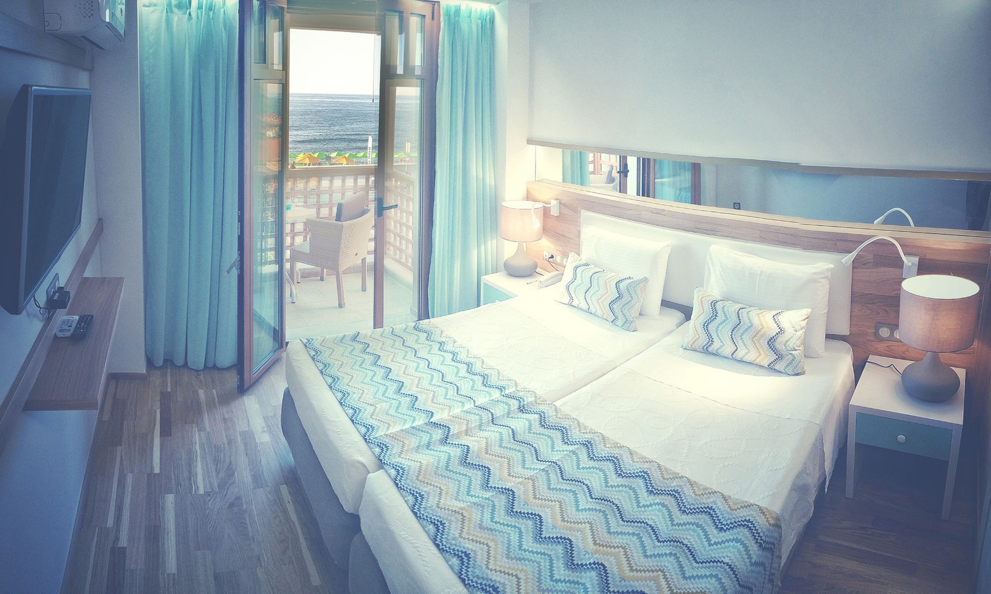 Quarto de hotel com uma cama grande, uma mesa de cabeceira com abajur, televisão e vista para o mar através de uma porta de vidro deslizante.