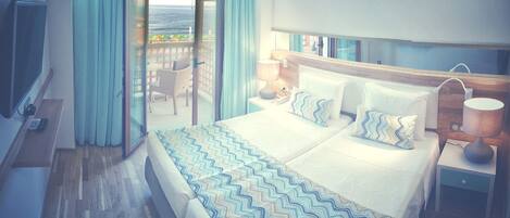 Quarto de hotel com uma cama grande, uma mesa de cabeceira com abajur, televisão e vista para o mar através de uma porta de vidro deslizante.