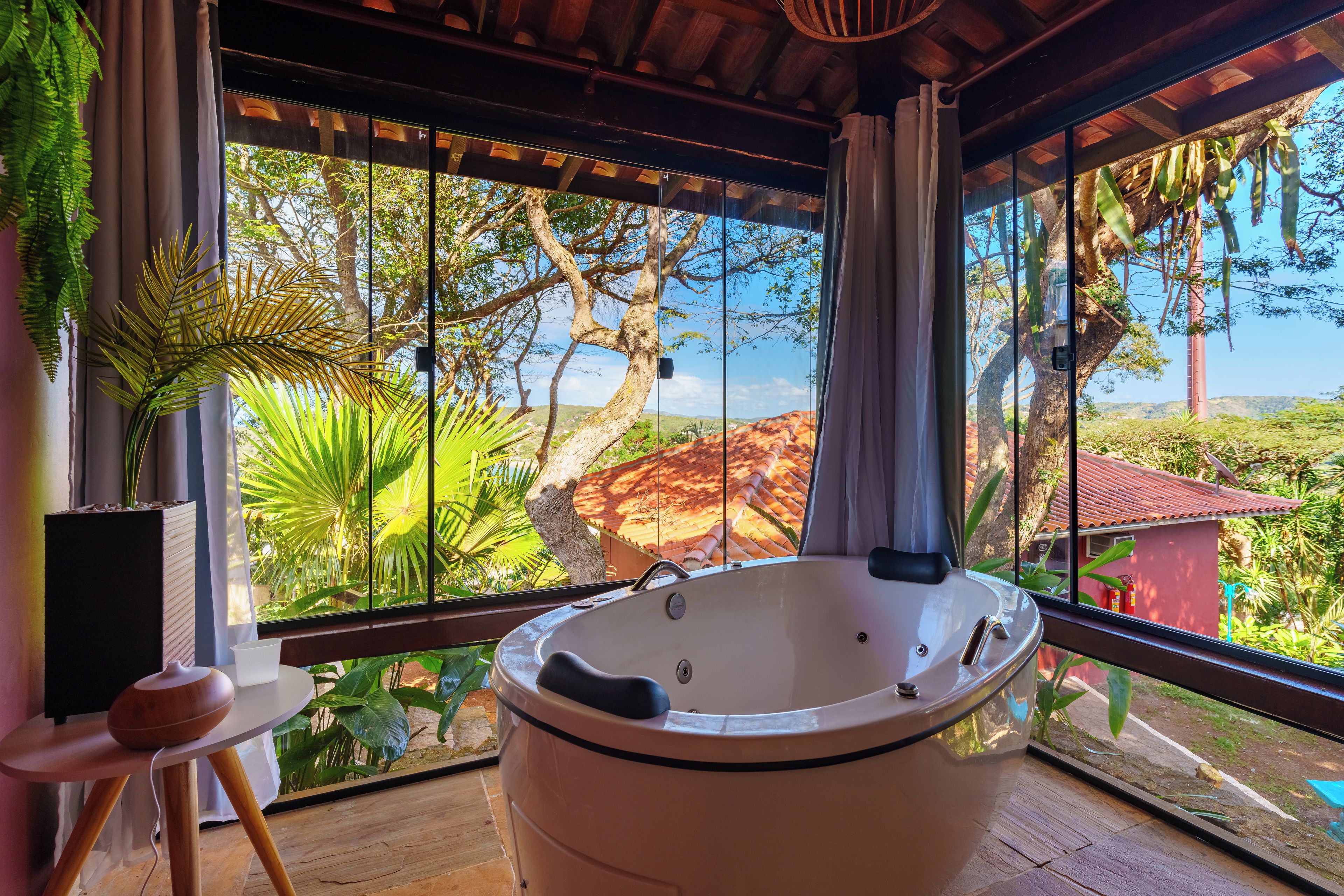 Un baño con una bañera grande, una mesita con un altavoz y vistas a un paisaje tropical a través de amplias ventanas.