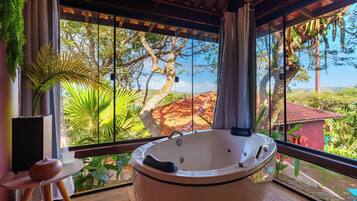 Un baño con una bañera grande, una mesita con un altavoz y vistas a un paisaje tropical a través de amplias ventanas.