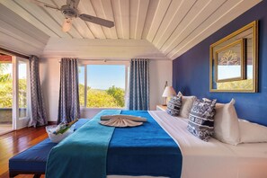 Quarto com cama, ventilador de teto, janela com cortinas e vista para o verde lá fora.