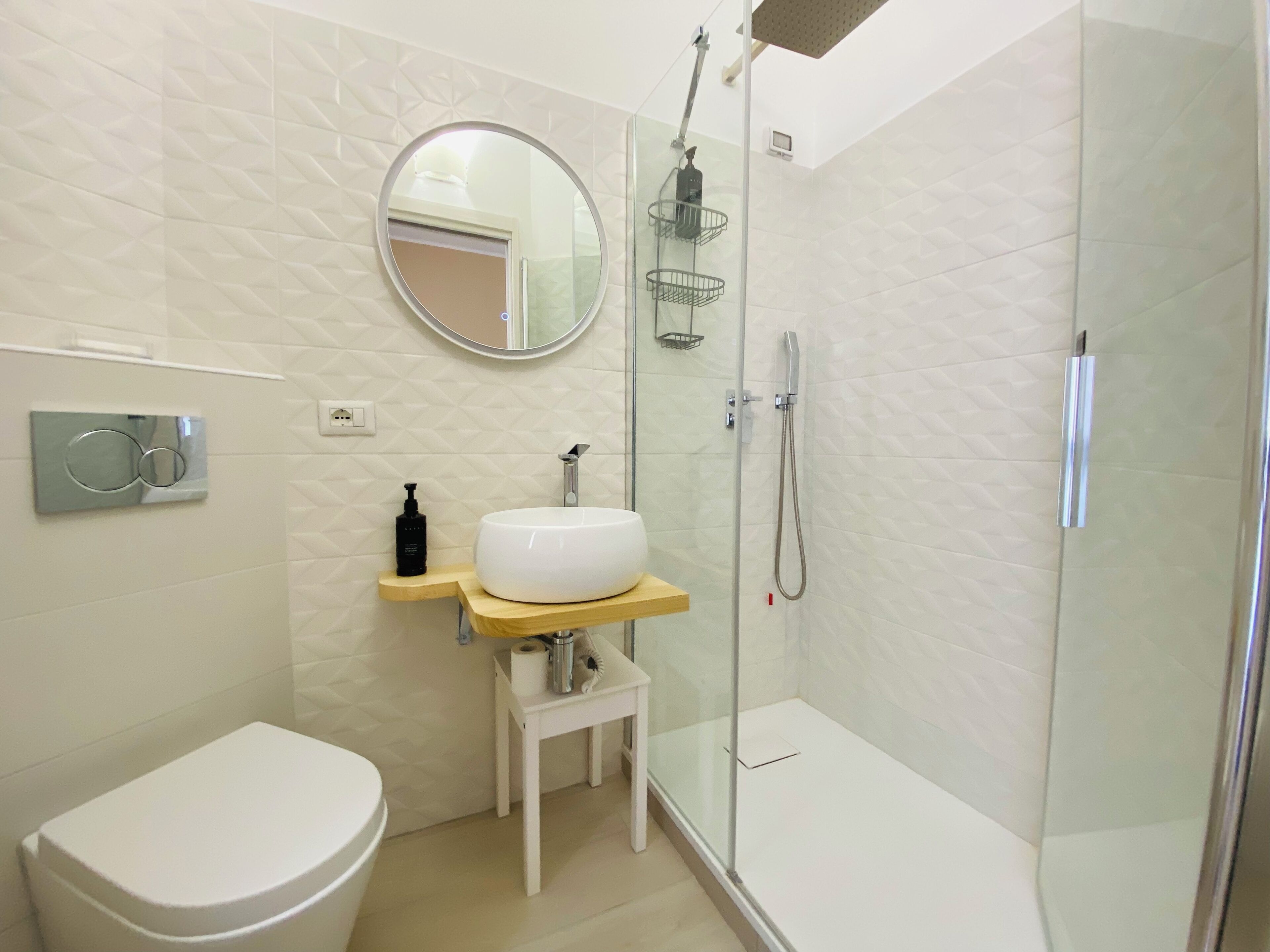 A modern bathroom with a toilet, a sink with a mirror above, a shower with a glass door, and a small table with toiletries.