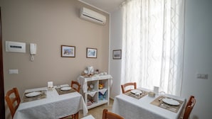A dining area with two tables, each set with plates, cutlery, and glasses. There are framed pictures on the wall, a shelf with books and decorative items, and an air conditioning unit above the tables. - Bed and Breakfast Dionisio (Taormina)