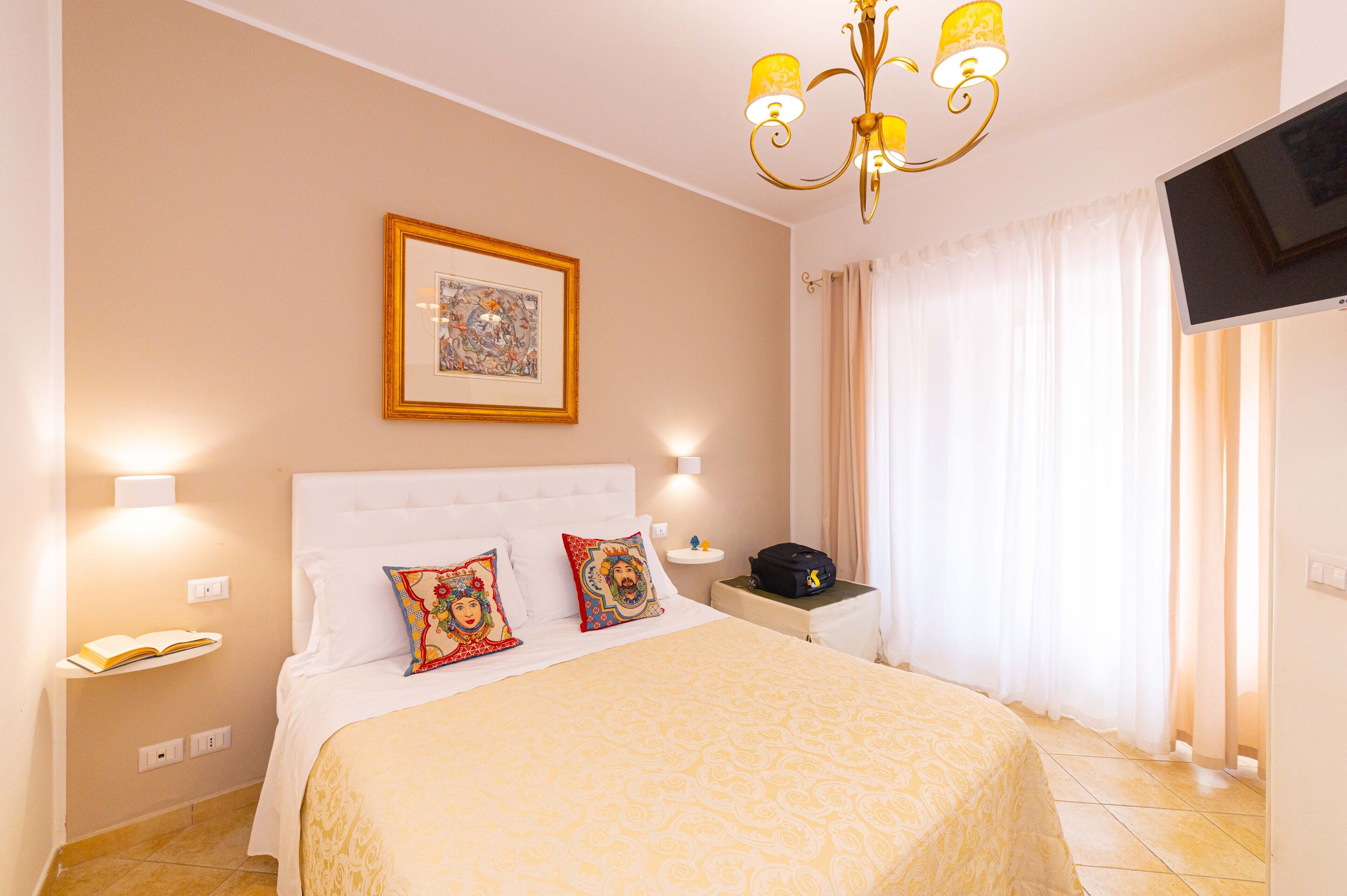 A hotel room with a bed, a bedside table, a TV, and a chandelier.