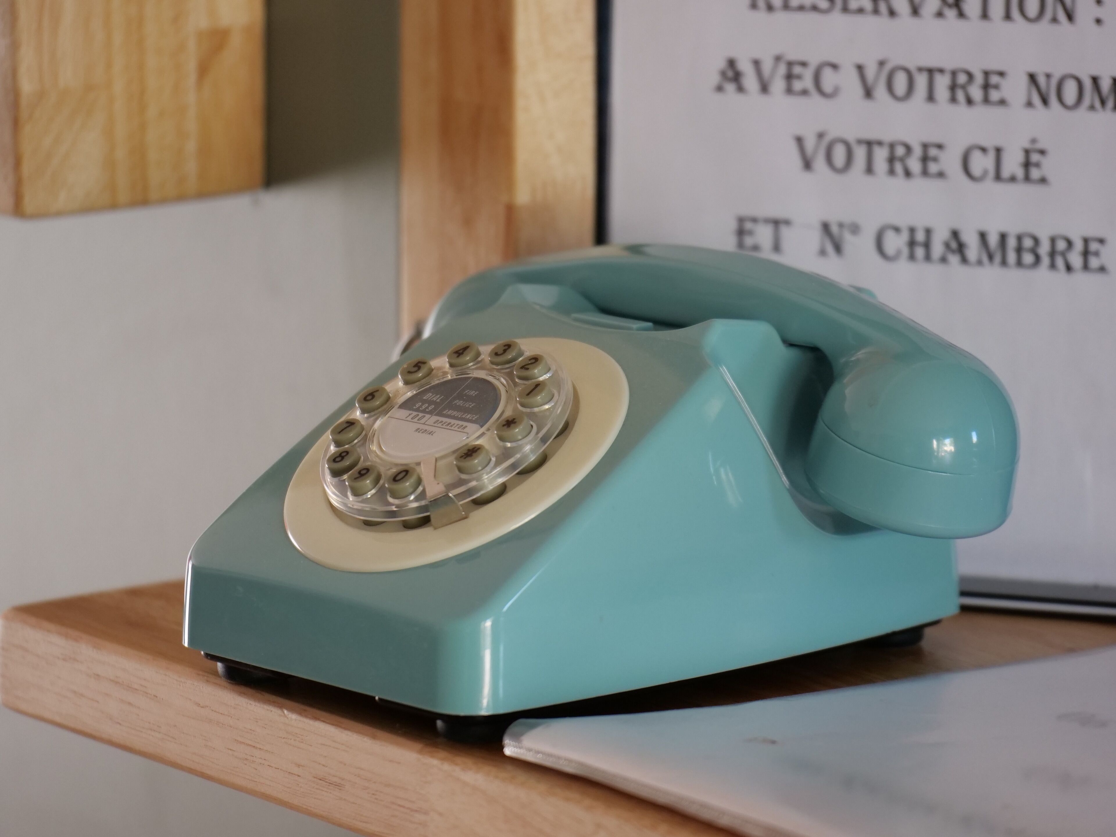 Un teléfono rotatorio color turquesa sobre una superficie de madera, con un cartel al fondo.
