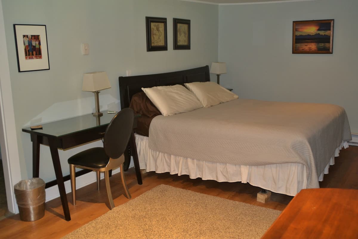 A bedroom with a bed, desk, chair, and a small table. There are two framed pictures on the wall, a lamp on the desk, and a picture of a sunset on the wall.
