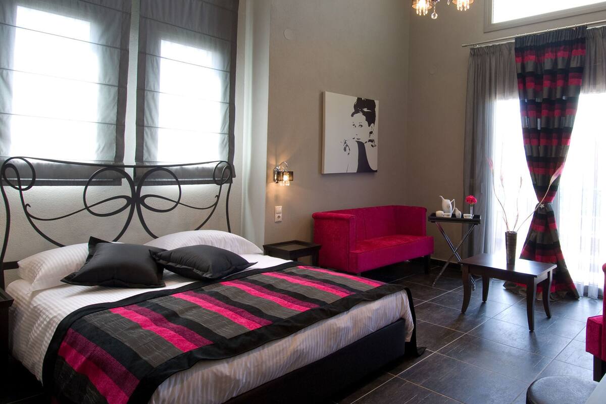 Un dormitorio con una cama, un sofá rojo, una mesita con un florero y un cuadro en la pared.