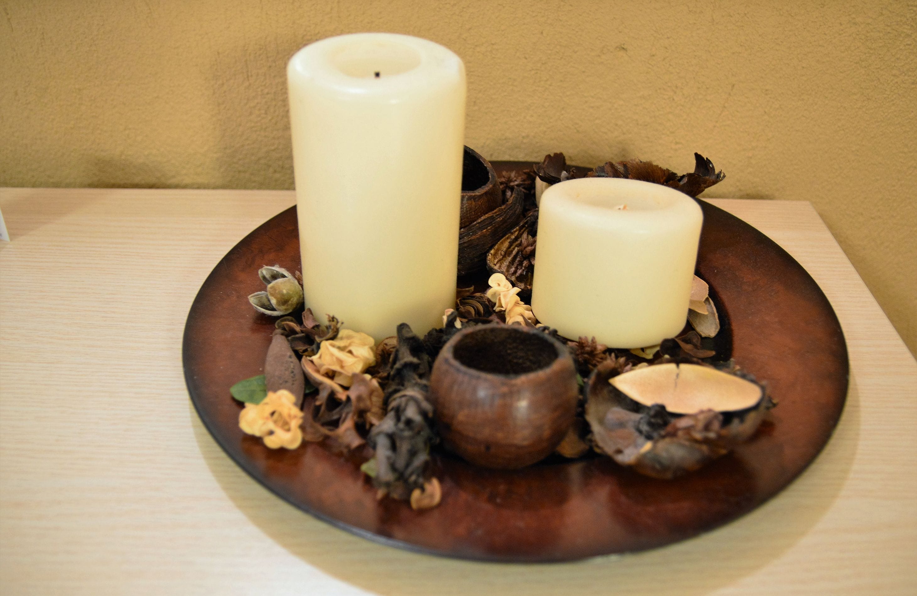 Two candles on a wooden plate with dried flowers and a small bowl.