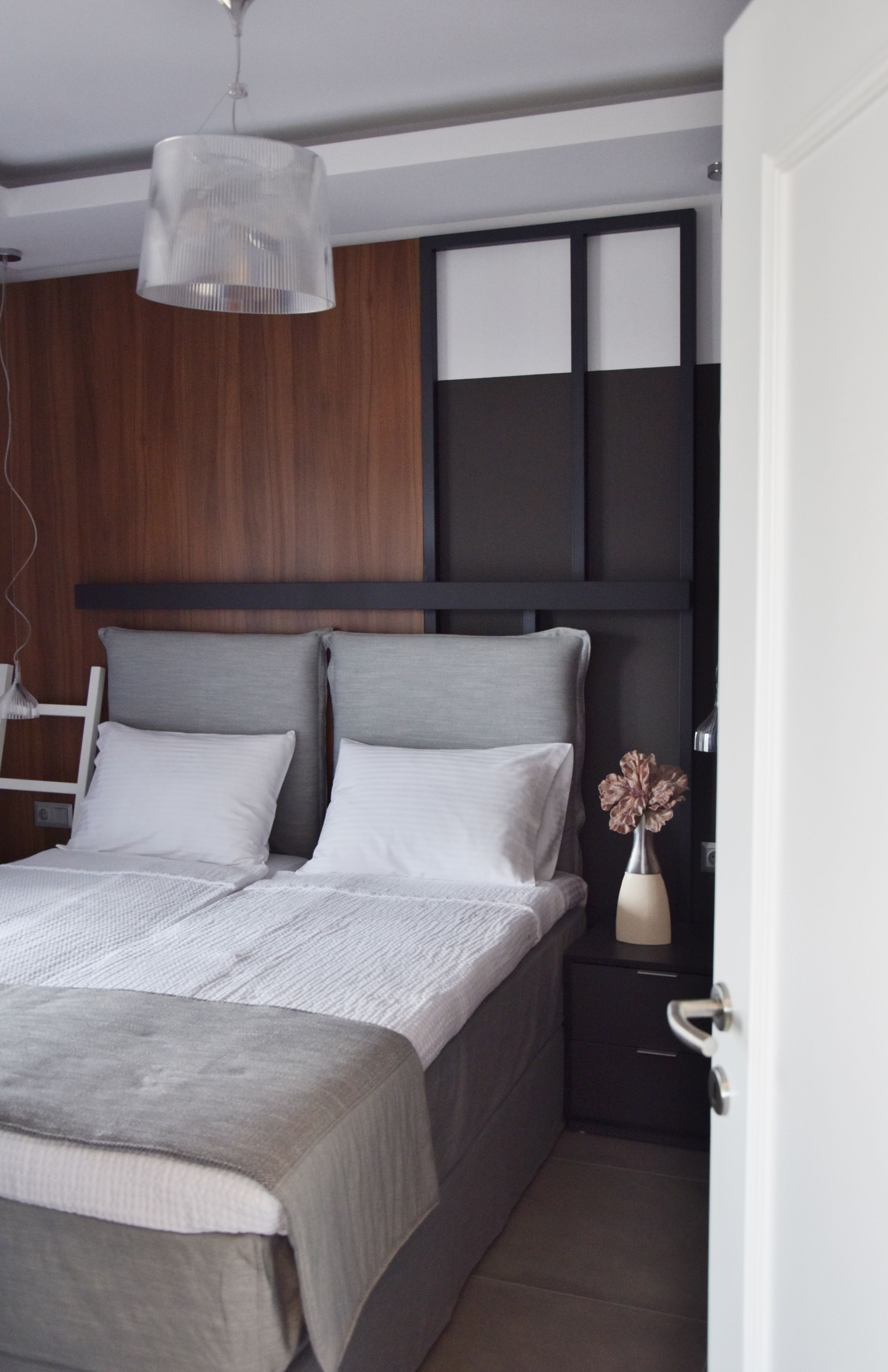 A modern bedroom with a large bed, a wooden headboard, a bedside table with a vase, and a hanging light fixture.