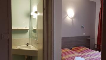 A hotel room with a bathroom featuring a sink, mirror, and a toilet. There is a bed with a checkered blanket and a towel on it.
