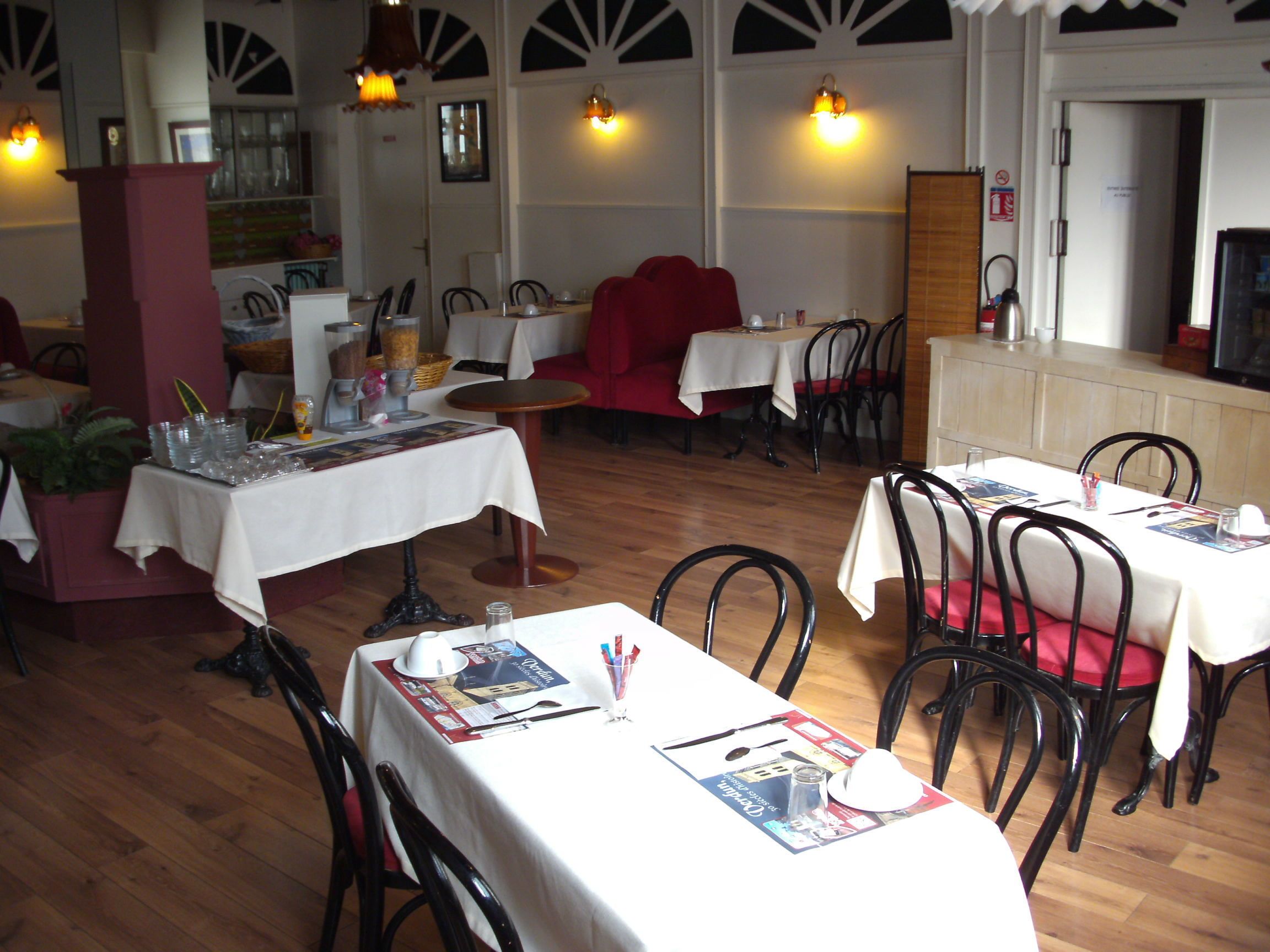 A restaurant with multiple tables, each covered with a white tablecloth. There are chairs around the tables, and some tables have menus and condiments on them. The restaurant has a wooden floor and a red couch in the background.