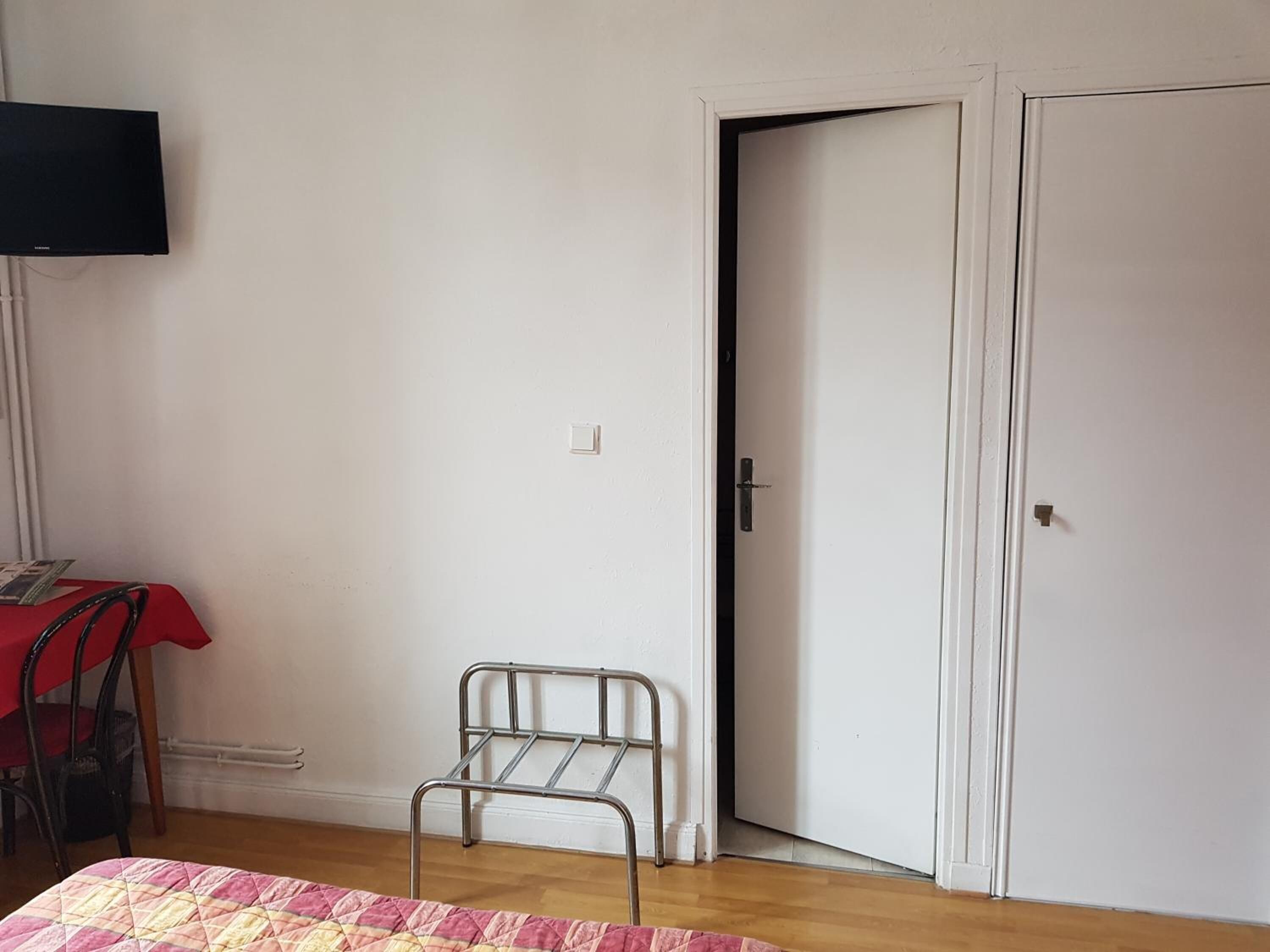A room with a bed, a metal chair, a table with a red tablecloth, a television mounted on the wall, and a door.