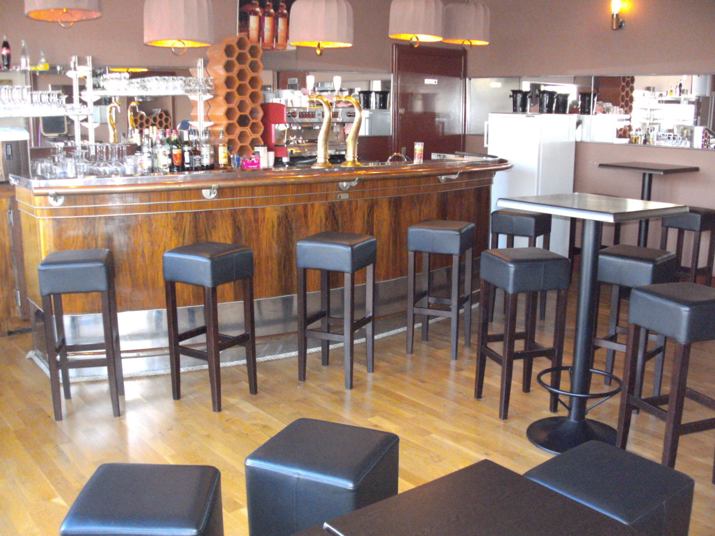 Un espace bar avec des tabourets en bois, un comptoir où sont exposées différentes bouteilles, et un coin salon avec des tables et des chaises.