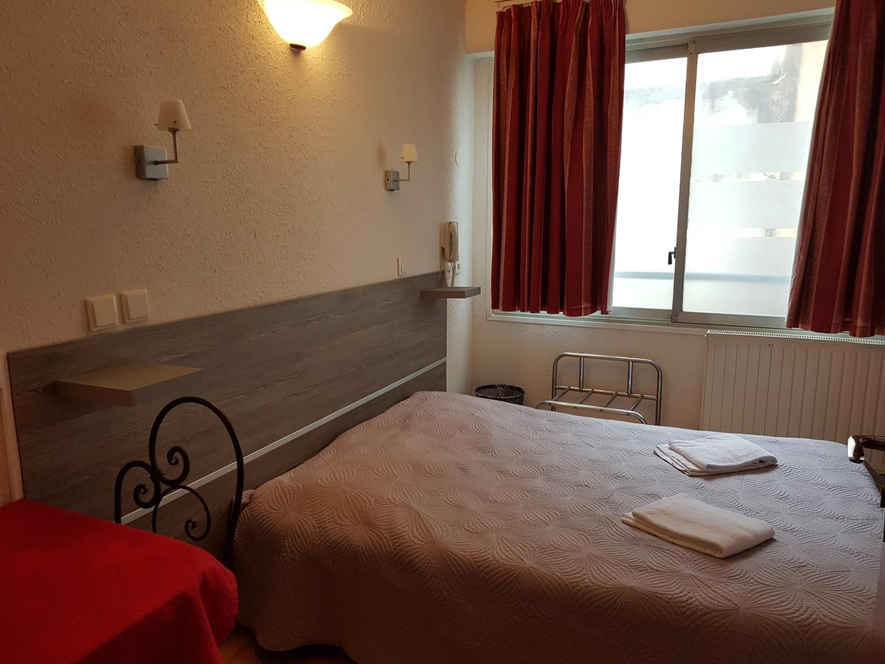 A hotel room with a bed, a bedside table, a wall-mounted lamp, a window with curtains, and a radiator.