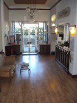 Un área de recepción con piso de madera, mostrador de recepción, zona de estar con sofá y una mesita, y una puerta de cristal que da al exterior.