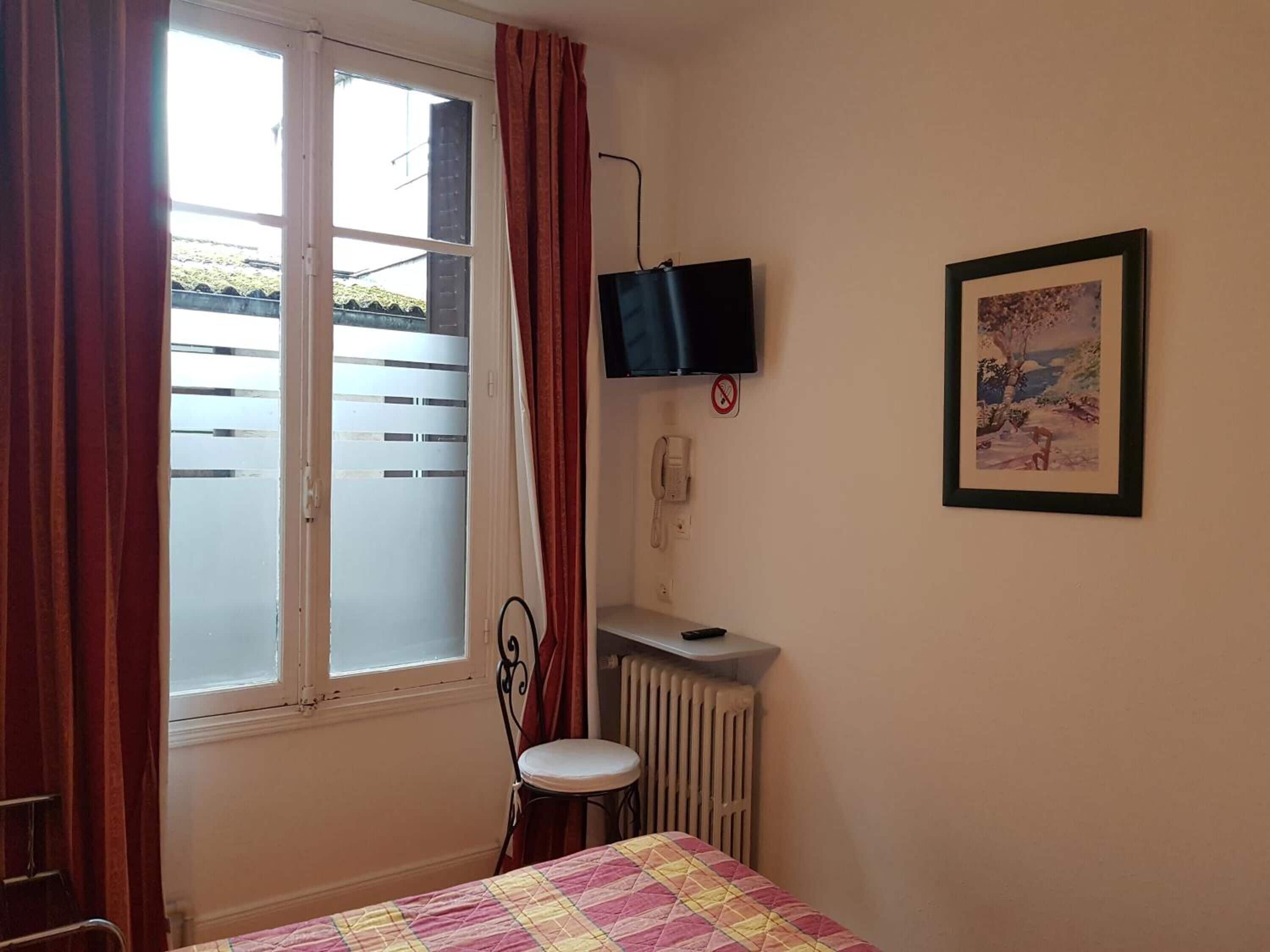 A bedroom with a bed, a window with curtains, a radiator, a desk with a phone, a television, and a framed picture on the wall.