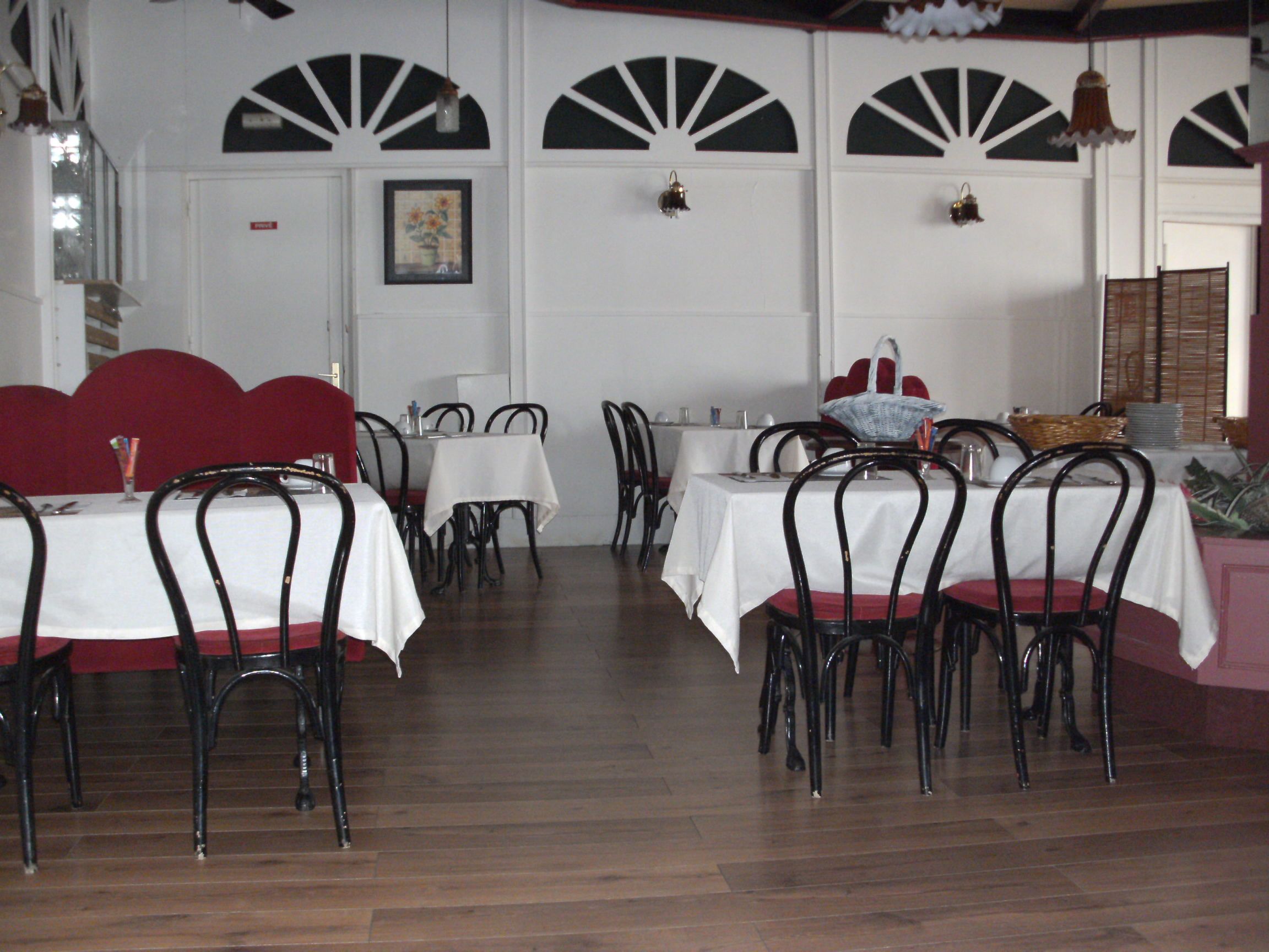 A restaurant interior with multiple tables and chairs, white tablecloths, and a wooden floor.