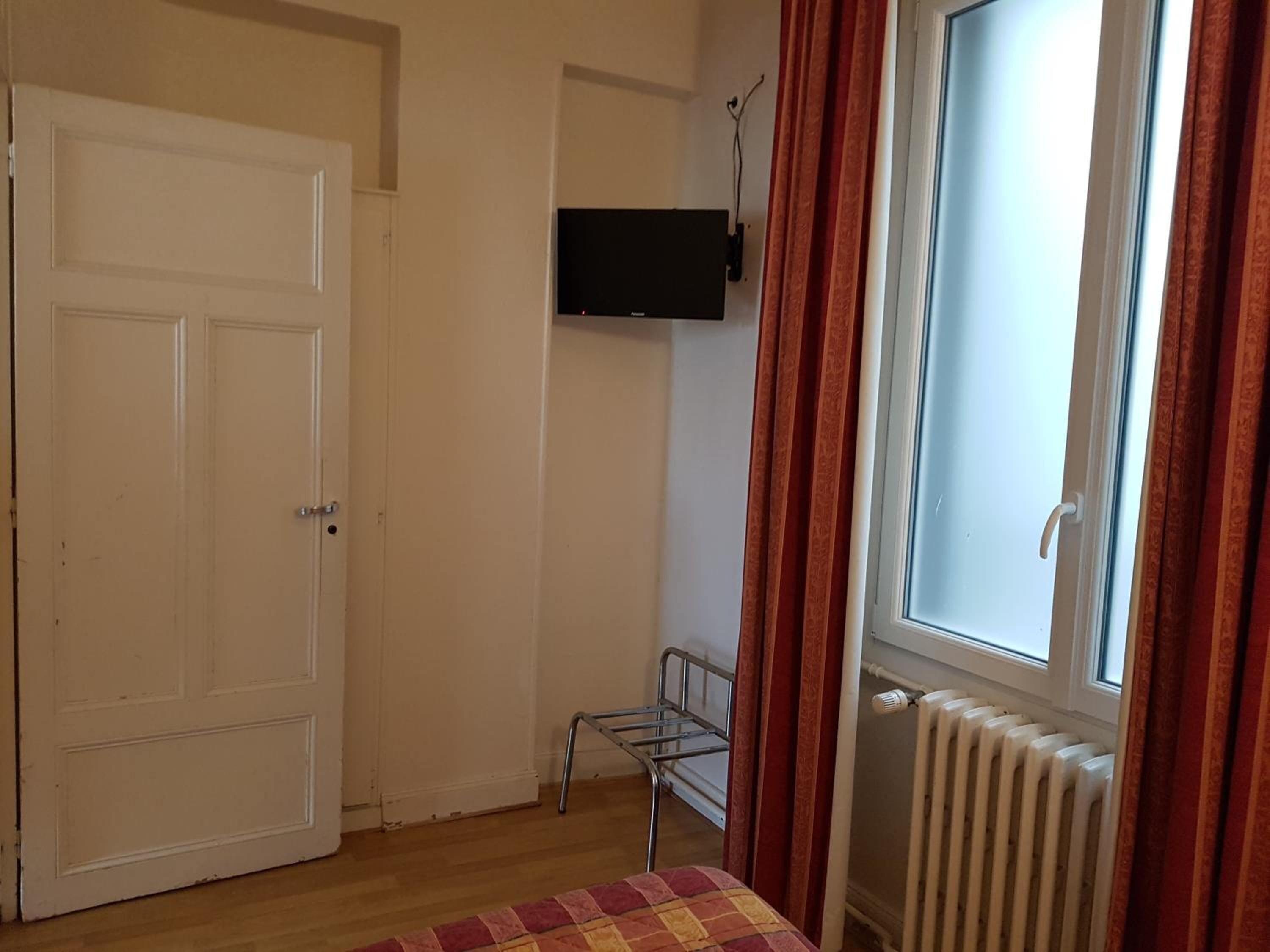 Una habitación con una puerta blanca, un radiador, una ventana con cortinas rojas y un televisor montado en la pared.