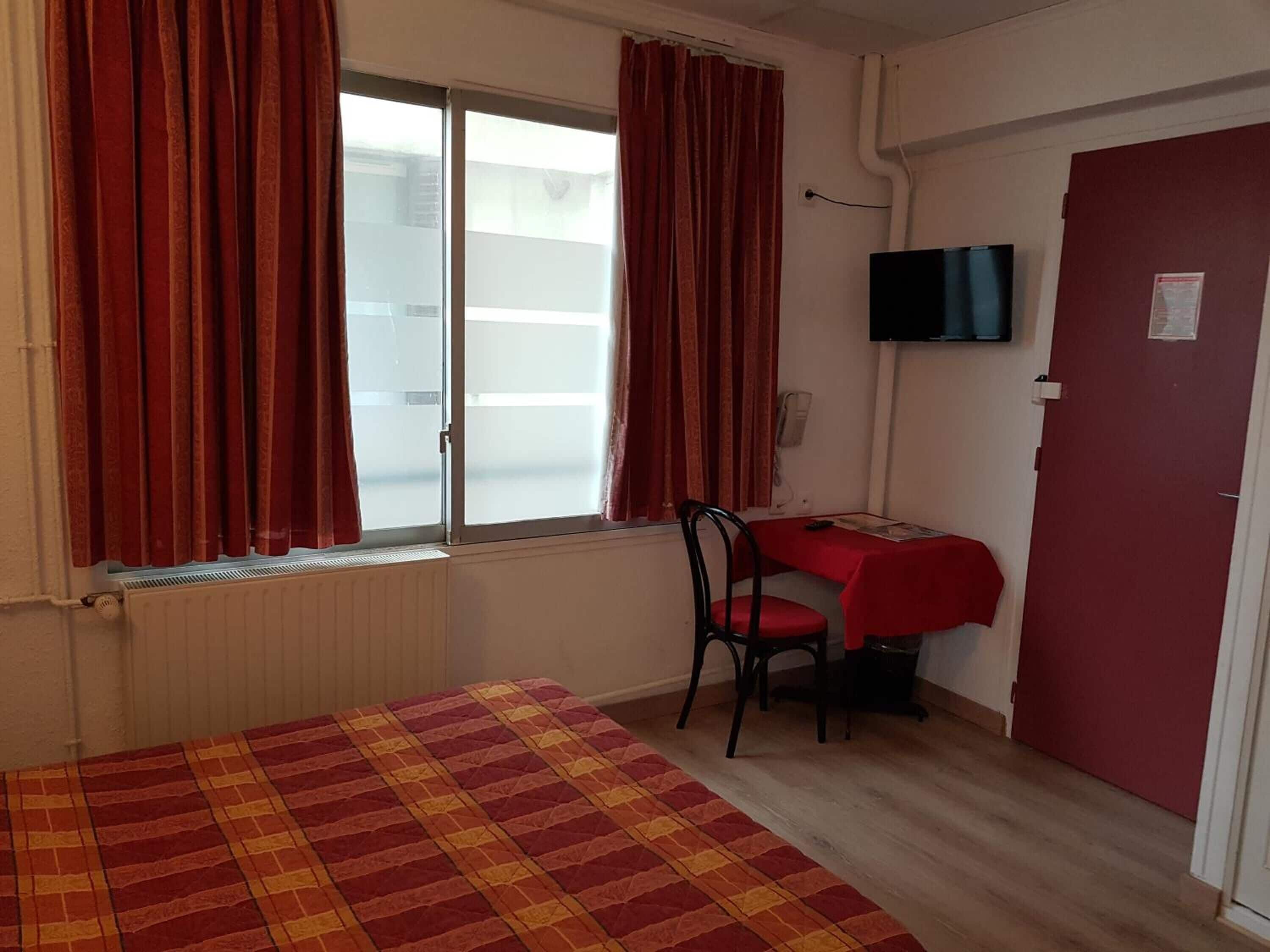 A small, tidy room with a bed, a desk, and a chair. There is a window with curtains, a door, and a TV mounted on the wall.