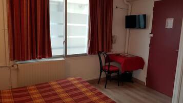 A small, tidy room with a bed, a desk, and a chair. There is a window with curtains, a door, and a TV mounted on the wall.