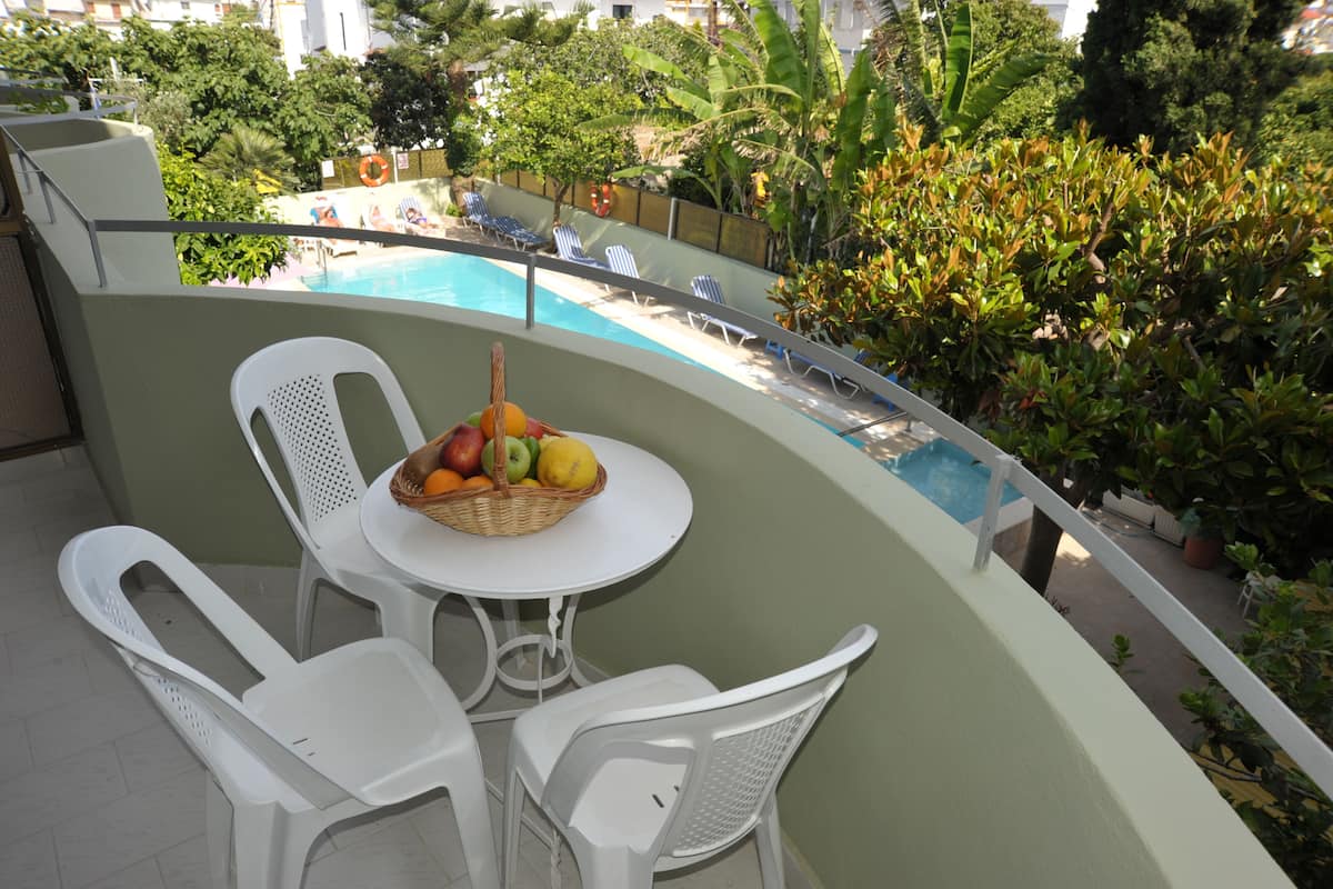 A balcony with a pool, white plastic chairs, a table with a fruit basket, and a view of the pool area.