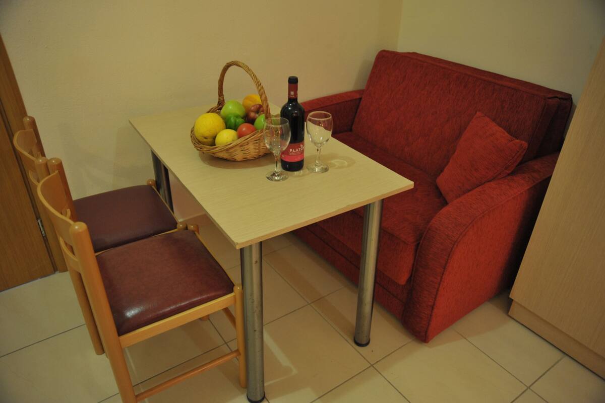 A small room with a red sofa, a wooden chair, a table with a basket of fruit and a bottle of wine, and two glasses.