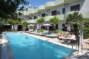A swimming pool with lounge chairs and umbrellas, surrounded by a building with multiple balconies and air conditioning units. - Anseli Hotel (Rhodes)