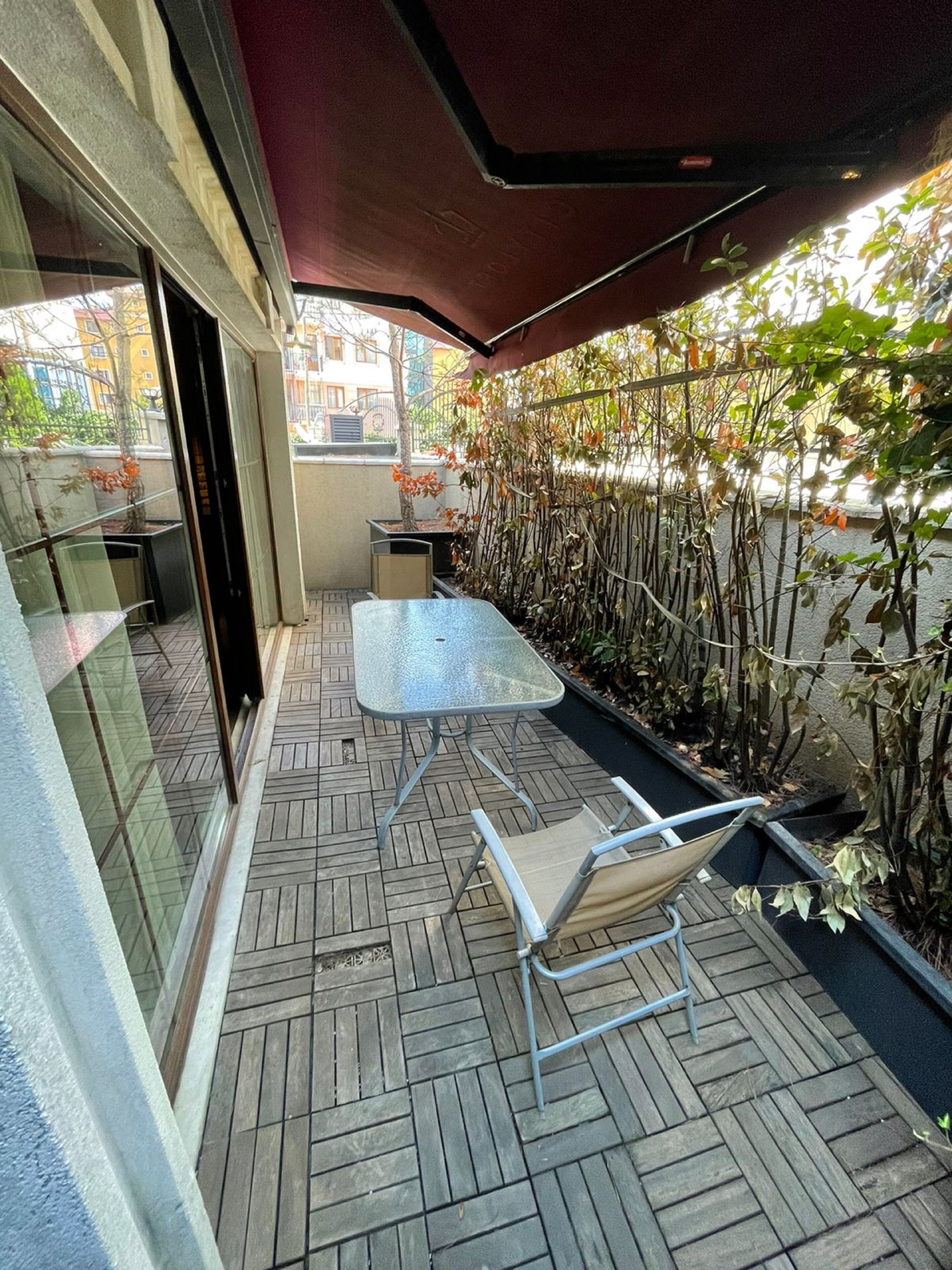 A covered patio with a table and chairs, a glass wall, and a wooden floor.